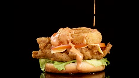 a close-up sequence shows thousand island sauce being poured over a fried chicken burger topped with crispy onion rings, shot against a black background with dramatic lighting