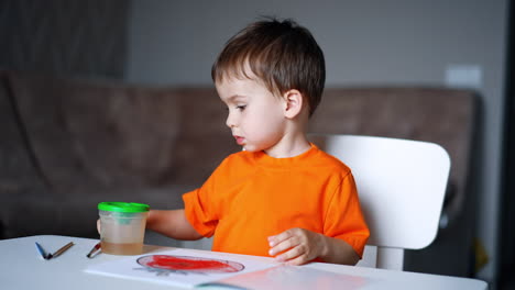cute toddler boy is preparing to paint. kid picks up a big brush and starts to paint on the sheet of paper. blurred room at backdrop.