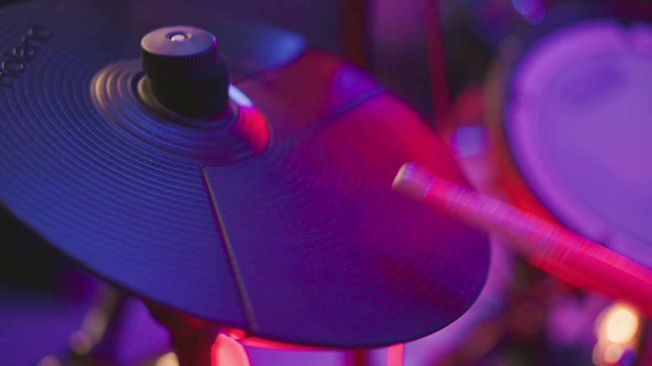 Close up of digital cymbal being struck by musician under vibrant studio lighting, showing motion and equipment detail during percussion practice with soft focus on surrounding background