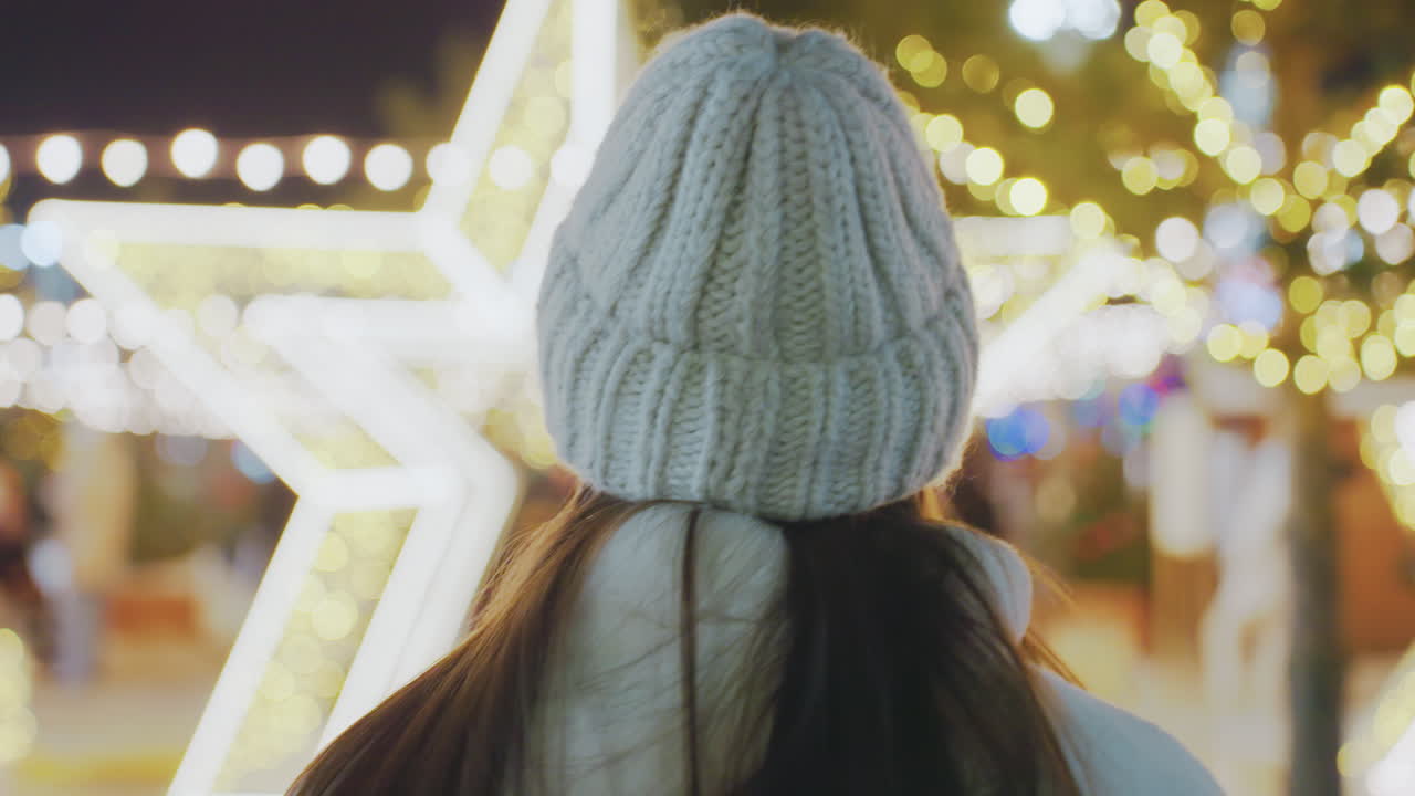 Close-up rear view of woman in warm winter outfit strolling through a beautifully illuminated city at night, star-shaped festive lights and blurred pedestrians