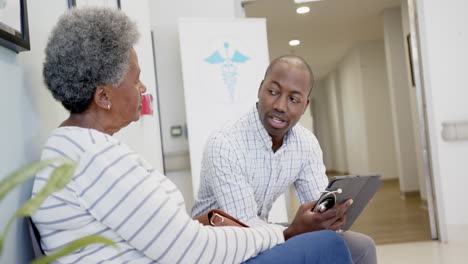 african american male doctor using tablet and talking with senior woman in hospital, slow motion