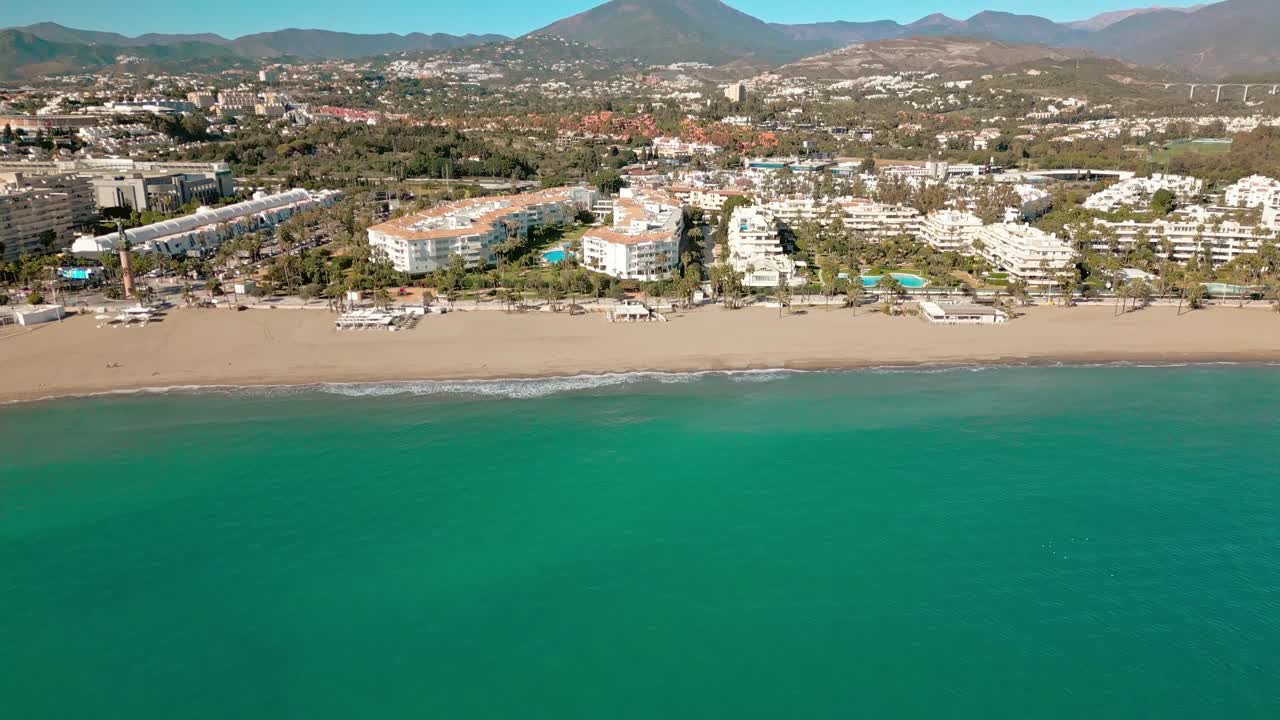 Ripresa drone inclinata dal mare verso Puerto Banús, resort sul lungomare di Marbella, che rivela la costa, la catena montuosa e il cielo azzurro della Costa del Sol, nel sud della Spagna