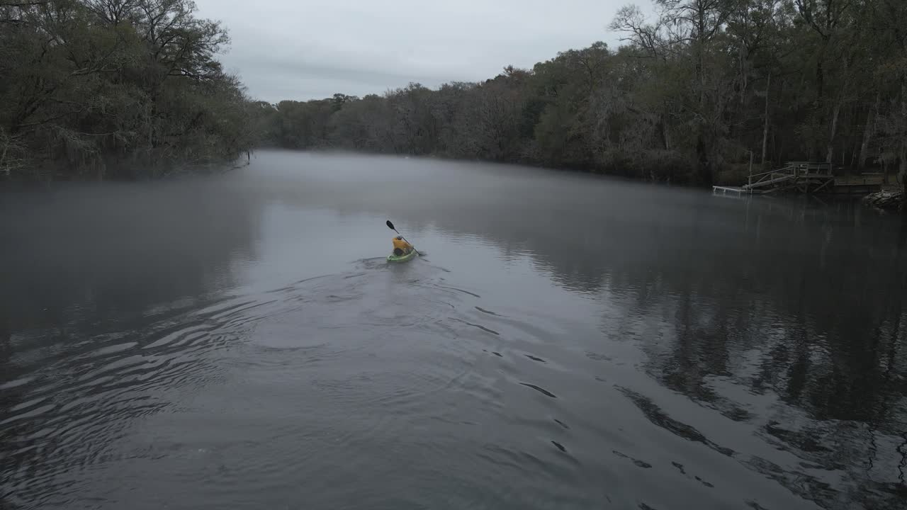 hombre remando un kayak en aguas oscuras cubiertas de niebla y rodeadas de árboles sin hojas grises