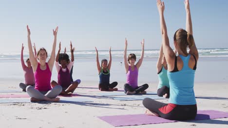 grupo multiétnico de mujeres haciendo yoga y poniendo los brazos en el aire en la playa y el fondo del cielo azul