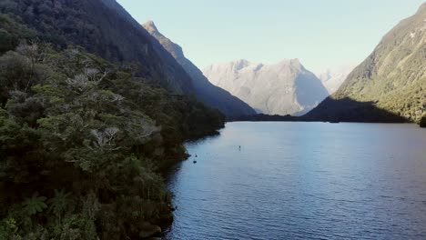 man paddle boards in remote mountain lake in fiordland national park in new zealand. think rainforest cloaks the lake.