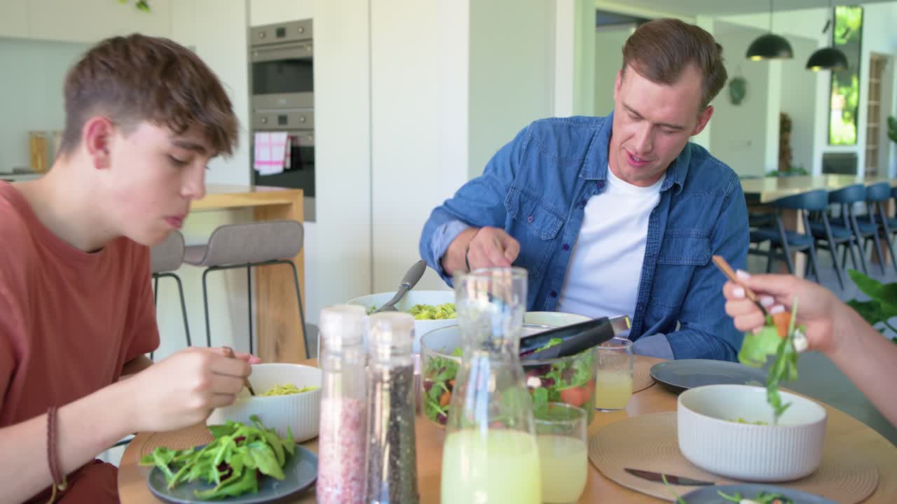 Family teen eating at kitchen table, man lifting pesto pasta from bowl, tasting for flavor coughing