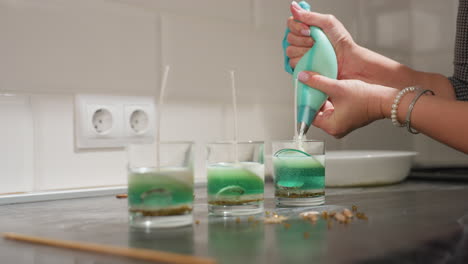 baker uses piping bag to fill green icing sugar into first glass container arranged on tabletop while preparing decorative layers in kitchen setup surrounded by tools and scattered ingredients