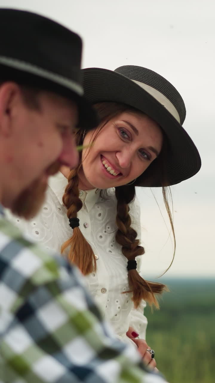 un artista con un sombrero y una camisa a cuadros está profundamente enfocado en pintar al aire libre. una mujer con un vestido blanco lo interrumpe juguetónamente, tocando suavemente su cara con una hoja de hierba