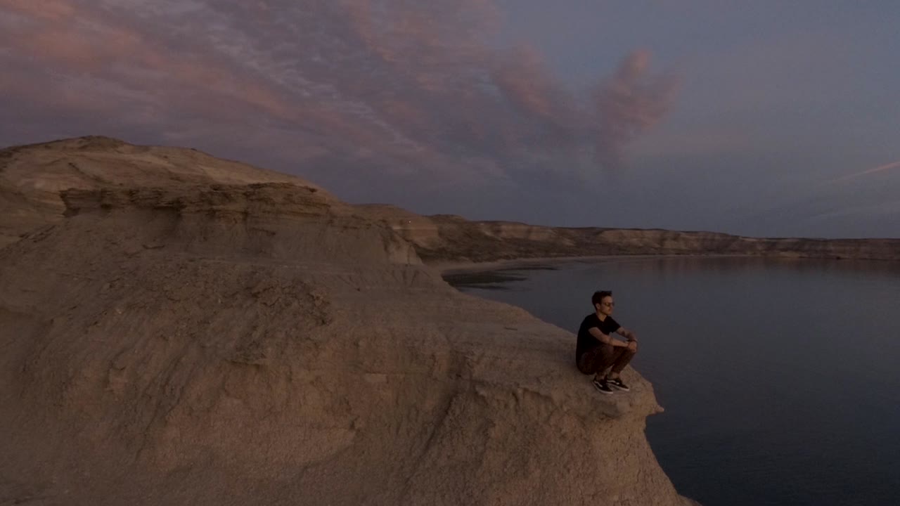 Giovane uomo solitario seduto sul bordo della montagna che guarda il mare in lontananza al tramonto - Ripresa aerea orbitale