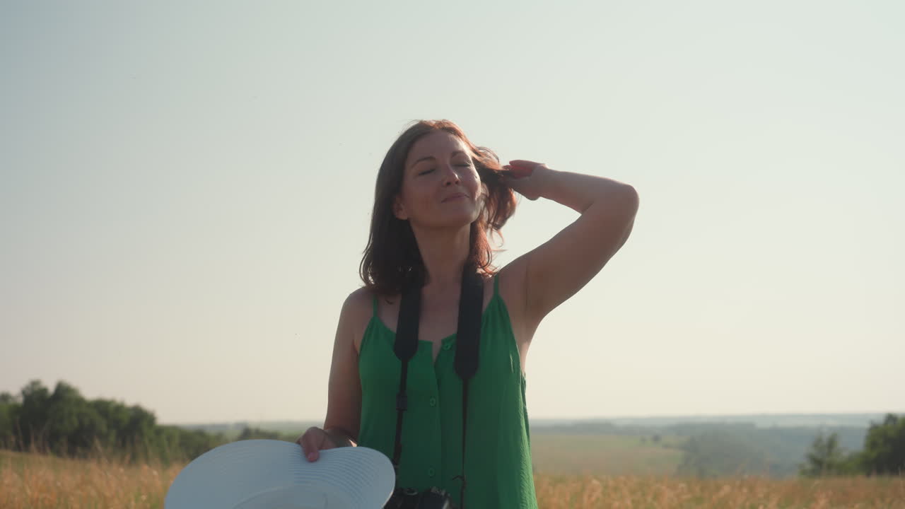 woman in green dress lifts sunhat from head while gently adjusting hair and placing hat back on under bright sunlight, standing in tall dry field with camera hanging from neck