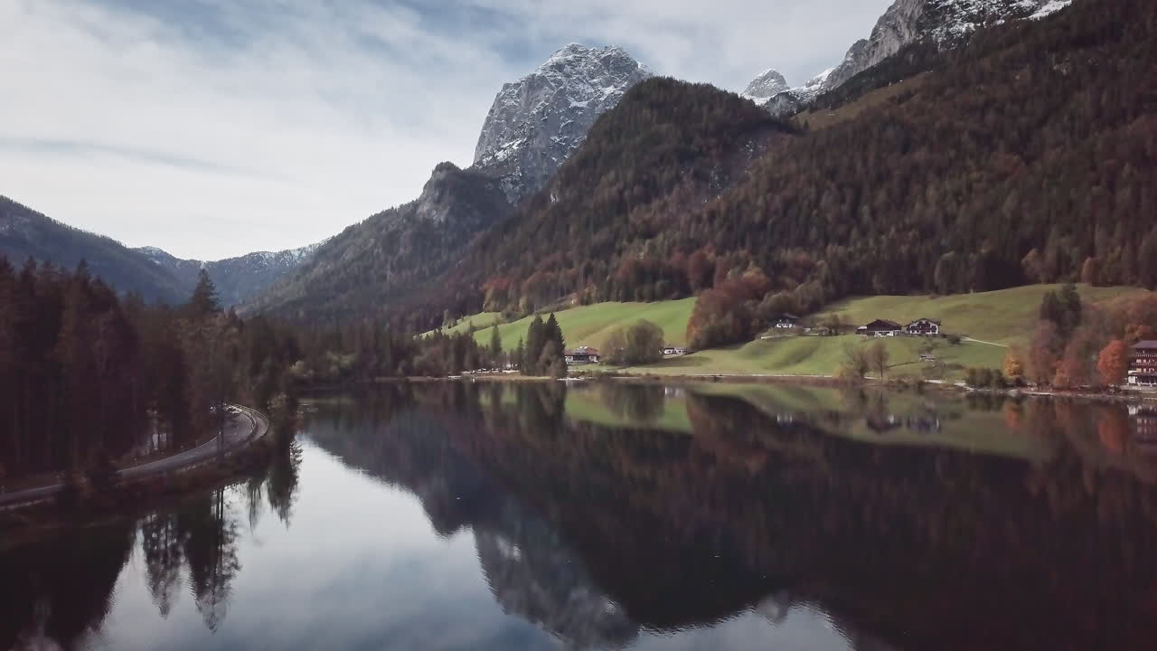 Un drone si muove in avanti appena sopra la superficie calma del lago Hintersee, scivolando verso prati verde brillante con chalet e imponenti montagne alpine innevate che si riflettono nell'acqua