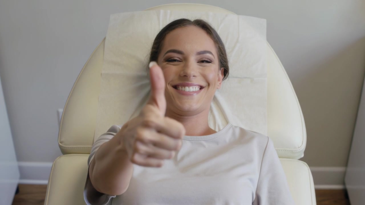 Mujer sonriendo a la cámara, recostada en una silla de clínica, levantando el brazo derecho y mostrando el pulgar hacia arriba, con una blusa clara.