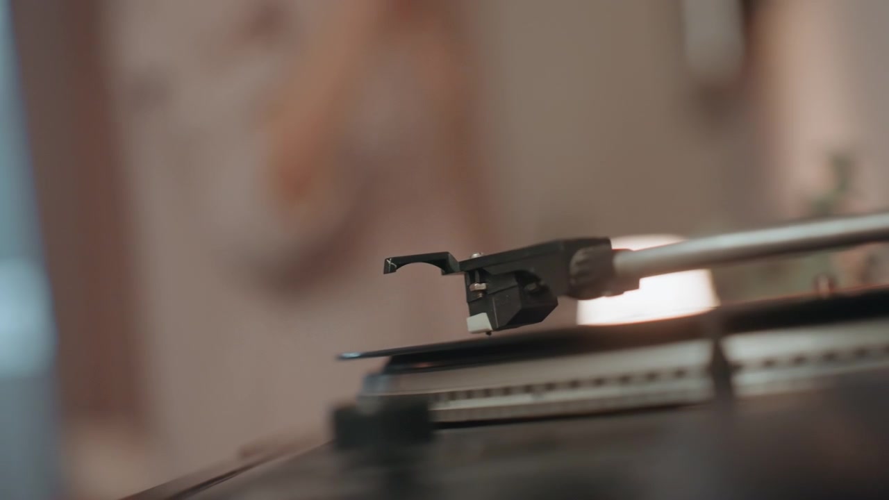 Vinyl record turntable in warm room, soft lamp bokeh and blurred figure in background, tonearm lowering stylus onto spinning record, intimate music lover listening to analog album, cinematic closeup