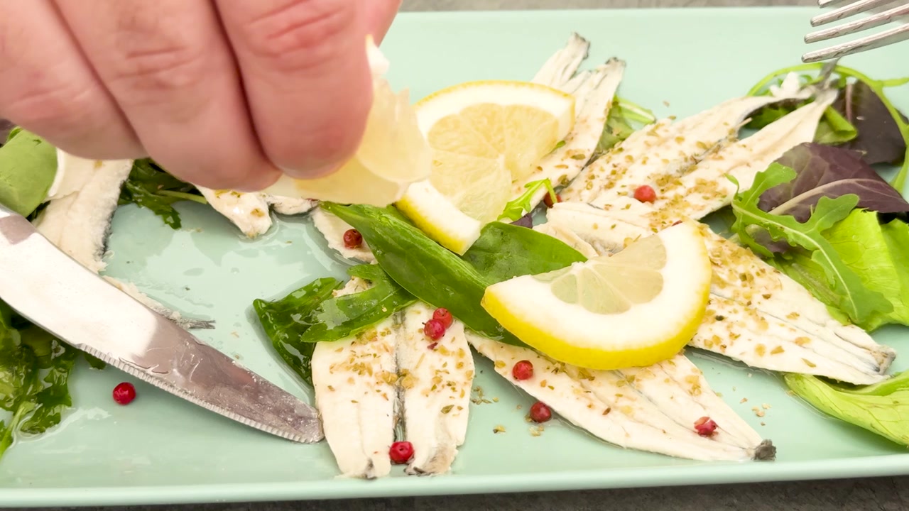 Nærbilde av en hånd som presser sitron over marinerte fiskefileter med urter og pepperkorn