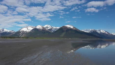 Drone-Se-Eleva-Sobre-Las-Aguas-Relucientes-Del-Brazo-Turnagain-En-Alaska-Con-Majestuosos-Picos-Nevados-De-Las-Montañas-Chugach-En-La-Península-De-Kenai-En-El-Fondo