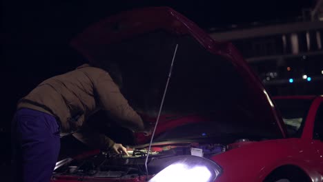 man looking at engine of broken down car with flashlight at night. headlights turned on