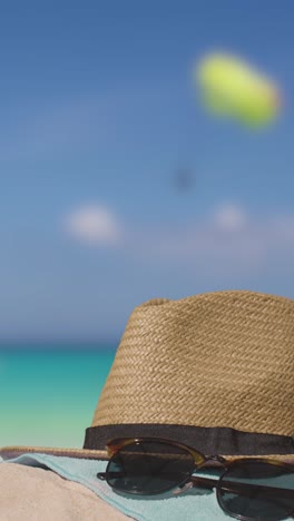 Vertical-Video-Summer-Holiday-Concept-Of-Sunglasses-Sun-Hat-Beach-Towel-On-Sand-Against-Sea-With-Paraglider