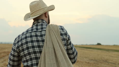 rückansicht des bauern mit hut und einem sack voller getreide auf dem feld