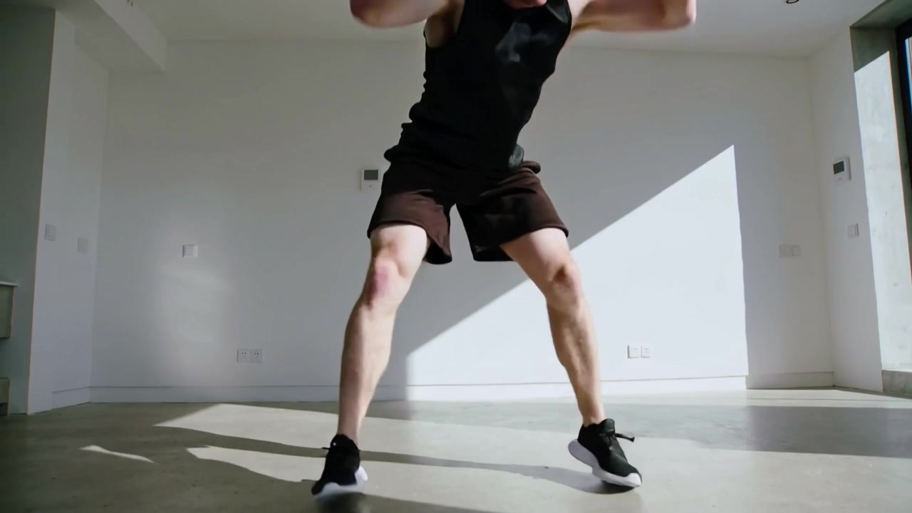 Uomo che inizia uno squat basso ed esegue burpees a casa, su una zona illuminata dal sole, con abbigliamento sportivo nero, in allenamento