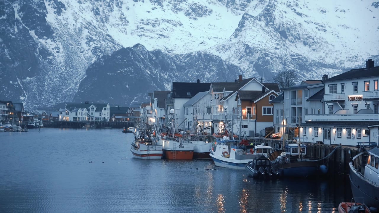 Henningsvær i Vågan, Nordland, Norge, vender mot Vestfjorden, med fiskebåter og bryggehus som gløder i blåtimen under Austvågøyas topper, og skaper et idyllisk postkortmotiv i et statisk bilde.