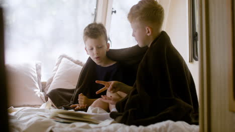 zwei kleine brüder mit decke auf den schultern sitzen in einem wohnmobil auf dem bett und spielen steinscherenpapier