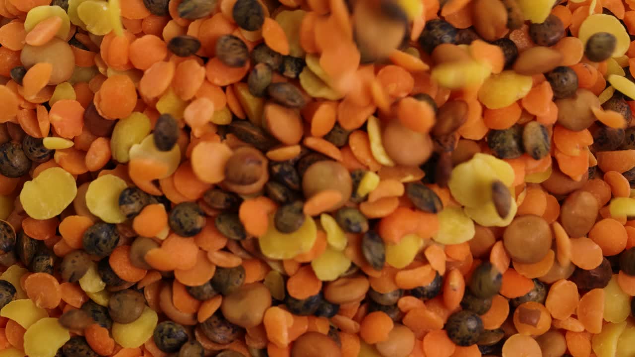 Vibrant lentils and chickpeas cascade onto a surface, creating a dynamic, colorful pattern in bright lighting