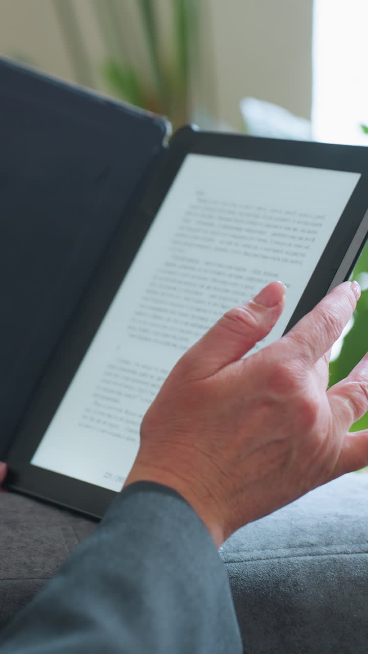Middle-aged woman reading digital book on e-reader while relaxing on gray couch surrounded by indoor plants in cozy living room environment during peaceful afternoon moment of solitude