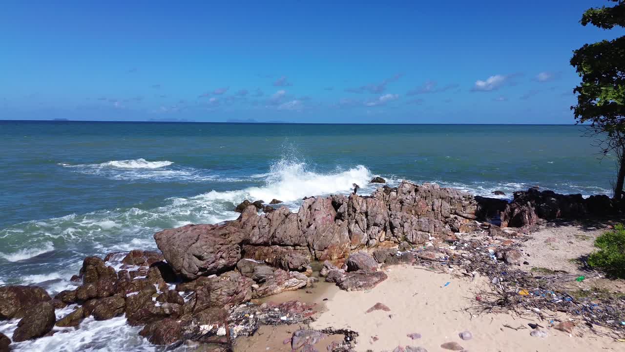 Aerial drone footage of a bird perched on rocky shoreline with waves crashing on the coast in Thailand, scenic seascape with wildlife and rugged tropical environment