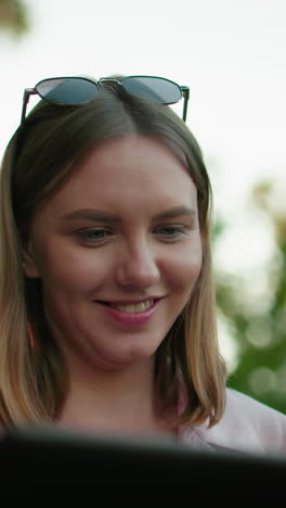 dama de camiseta rosa con gafas de sol descansando en la cabeza, sonriendo cálidamente mientras se centra en algo delante de ella, el fondo presenta árboles con la luz del sol filtrándose a través