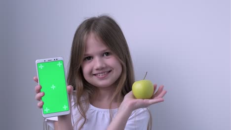 cute child girl shows apple and phone with green screen mock up cellphone, mobile, telephone. chroma key green screen smartphone for advertising. fruits, healthy eating, dental care for children