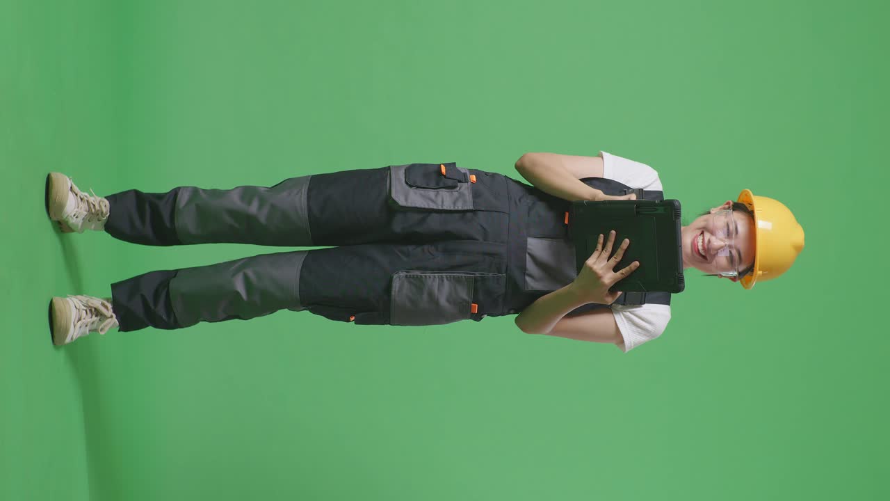 полное тело азиатской женщины рабочая в защитных очках и защитном шлеме с помощью планшета, стоя в зеленом экране фоновой студии