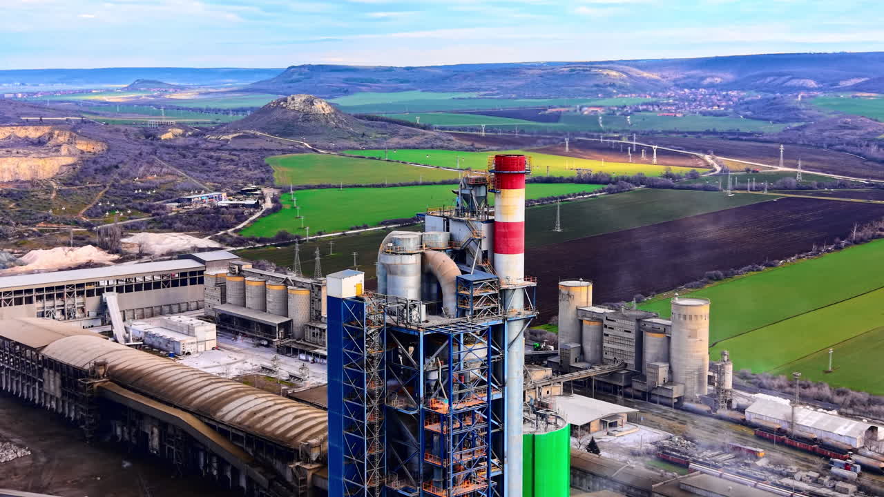 Vista aerea di uno stabilimento di produzione con alte ciminiere e silos che si affaccia su verdi colline e campi agricoli. Ciminiera industriale in paesaggio rurale.