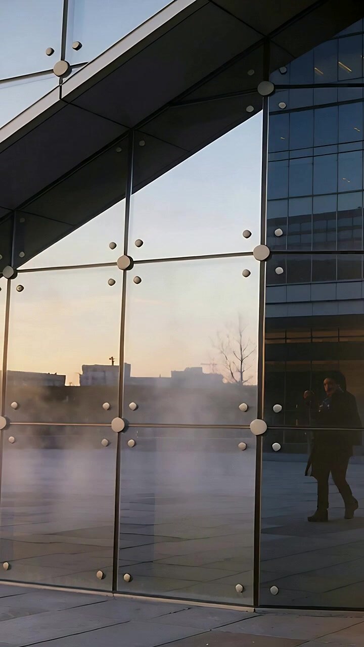 Video vertical: Peatón caminando que aparece a través de una pared de cristal en una plaza al amanecer, vistiendo un abrigo oscuro