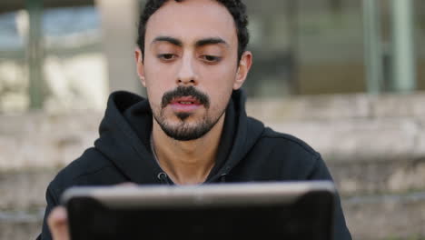 Front-view-of-young-Arabic-handsome-man-with-dark-curly-hair-and-beard-in-black-hoodie-sitting-on-stairs-outside