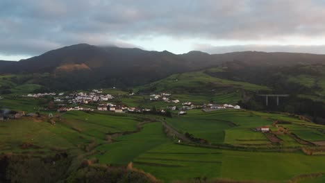 Drone-footage-of-lush-green-volcanic-island-countryside-at-sunset-with-homes-and-road-near-ocean-cliff-and-fog-covered-mountain-in-backdrop