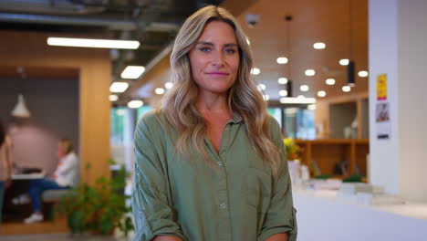 Portrait-Of-Mature-Businesswoman-Standing-In-Busy-Open-Plan-Office-With-Colleagues-In-Background