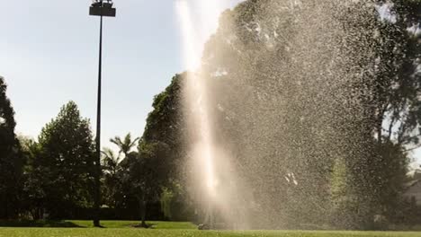 Ein-Rotierender-Sprinkler-Spritzt-Wasser-Durch-Den-Park,-Im-Hintergrund-Die-Parklicht-Silhouette