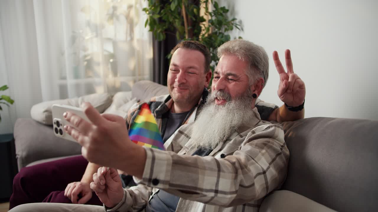hombre mayor feliz con color de cabello gris y exuberante barba blanca en una camisa a cuadros sostiene una bandera lgbt y toma una selfie usando un teléfono blanco junto con su novio moreno en un sofá gris en un apartamento moderno
