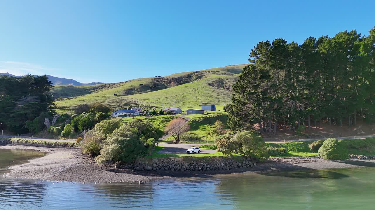 Aerial footage captures Akaroa's serene lake, lush greenery, and rolling hills under clear blue skies