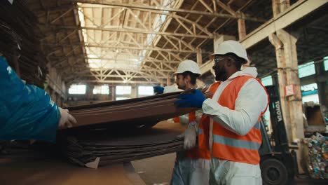 Ein-Trio-Von-Menschen-In-Weißen-Schutzuniformen-Und-Orangefarbenen-Westen-Sortiert-Riesige-Stapel-Altpapier-In-Einer-Abfallrecyclinganlage-Und-Lagert-Sie.-Zwei-Männer-Nehmen-Riesige-Pappbögen-Zusammen-Und-Bewegen-Sie