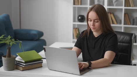 una mujer joven se comunica por chat en internet en una computadora portátil escribiendo y enviando mensajes sentada en una habitación de oficina moderna con un interior acogedor