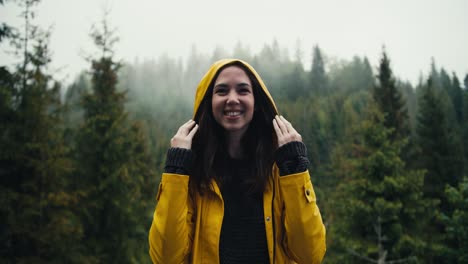 a happy girl in a yellow jacket stands against the backdrop of a coniferous mountain forest, she puts on a hood and looks into the camera. it's raining a little