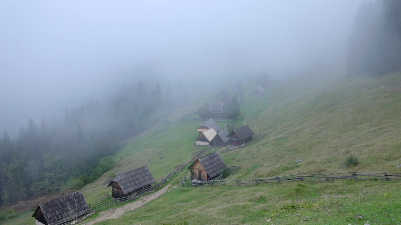 Dichter Nebel zieht über ein traditionelles Bergdorf mit Holzhütten. Gespenstische und atmosphärische Szenerie in einer ländlichen Landschaft an einem bewölkten Tag.