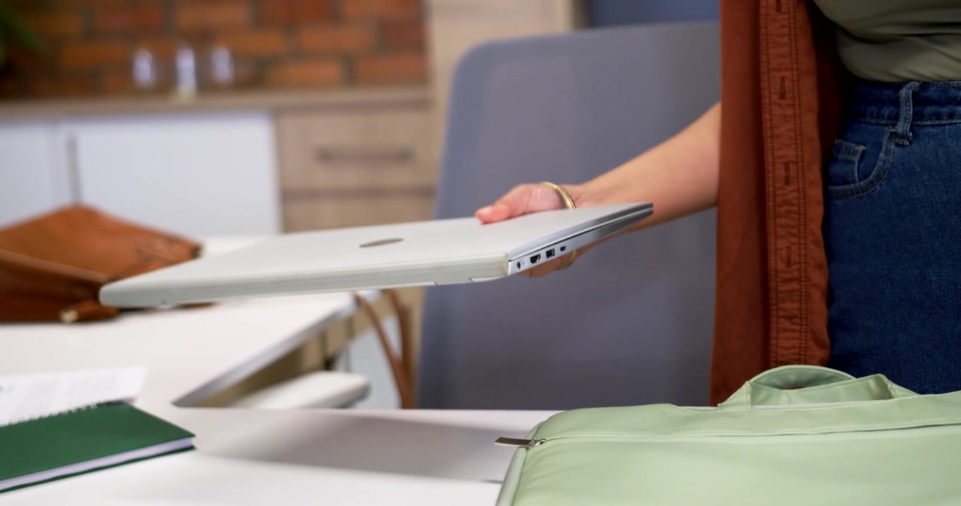 African American woman in cardigan at table opening green case, placing silver laptop for travel