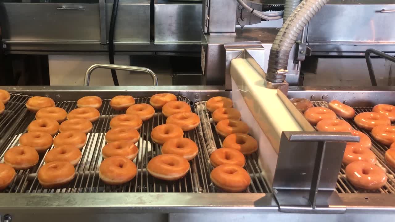 Hundreds of fresh ring doughnuts being glazed by a continuous wall of liquid sugar