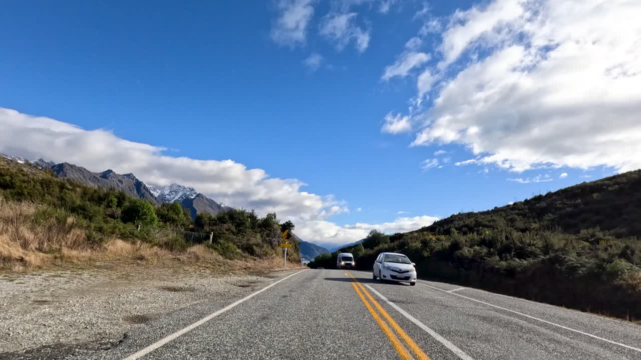 Vehicles travel along a picturesque mountain road in Queenstown, New Zealand, under clear blue skies with scattered clouds