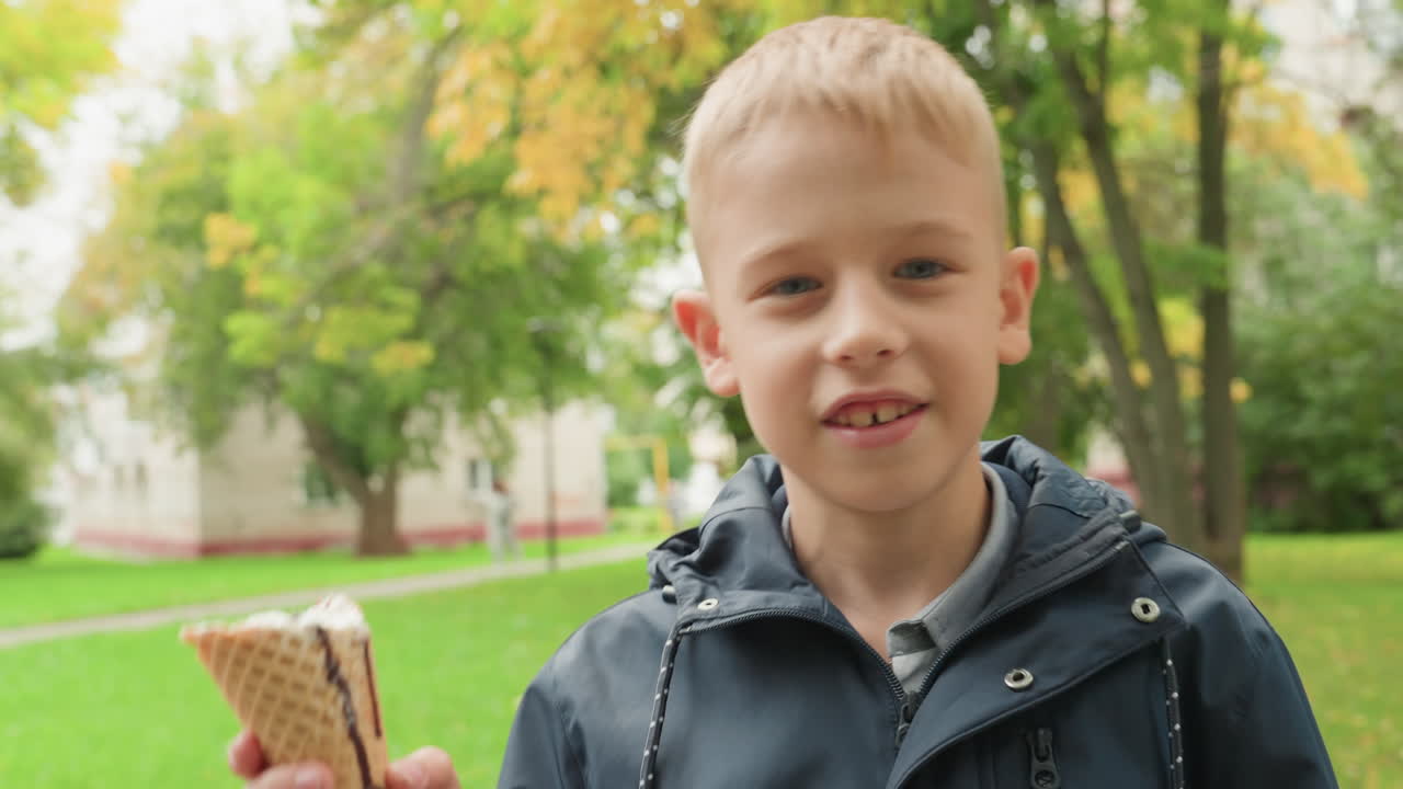 Ragazzo bianco al parco si prepara per il ritorno a scuola, gusta uno snack con cono, indossa una giacca con cappuccio, alberi autunnali, prato verde, primo piano spontaneo, sorriso timido, espressione curiosa, luce naturale soffusa, routine mattutina.