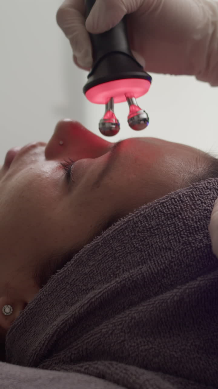 Kosmetolog bruger massageudstyr med rødt lys på ung kvindes ansigt