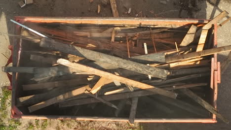 top-down view of an excavator arm loading long wooden beams into a dumpster as part of a demolition site cleanup process