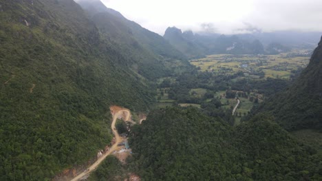 Drone-moves-over-windy-road-along-mountain-peak-to-reveal-a-small-village-at-the-valley-with-yellow-rice-field-at-the-background-Cao-Bang-Viet-Nam